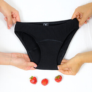 Vier Hände halten eine schwarze Periodenunterhose auf weißem Hintergrund, darunter liegen drei Erdbeeren